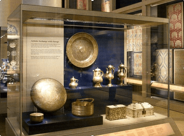 A display case with a blue background. A round platter is hanging on the back. In front of this, on a narrow shelf, are four coffee pots. In the foreground of the case are two small decorated caskets and some other decorative items in metal.