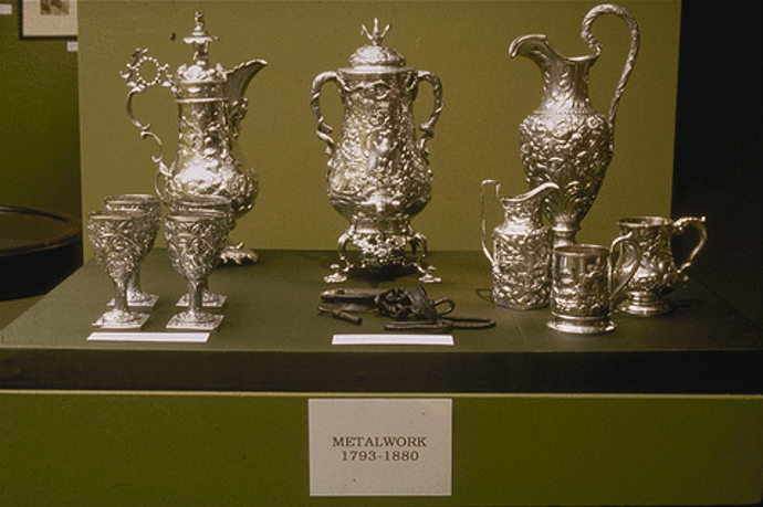 A display cabinet showing some highly ornate and decorated silverware. At the back three pitchers, at the front left five goblets and at the front right, a group of three small jugs. At the front in the middle, between the goblets and the jugs are a set of shackles. A label at the front of the display case reads "Metalwork 1793-1880".